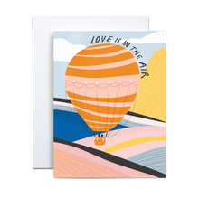 Load image into Gallery viewer, bold and abstract pink, yellow, cobalt, baby blue, black, and whitemountain and valley landscape with various textures and a pink and orange hot air ballon with two black shapes in the basket with 'love is in the air' text circling the balloon greeting card