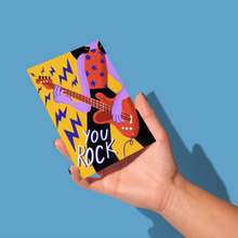 Load image into Gallery viewer, hand holding purple abstract person with red tank top and purple stars wearing yellow hoop earrings and black pants playing a red electric guitar with purple lightning bolts coming out with the text &#39;you rock&#39; on lower left side on a yellow background greeting card
