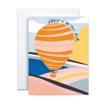 Load image into Gallery viewer, bold and abstract pink, yellow, cobalt, baby blue, black, and whitemountain and valley landscape with various textures and a pink and orange hot air ballon with two black shapes in the basket with &#39;love is in the air&#39; text circling the balloon greeting card
