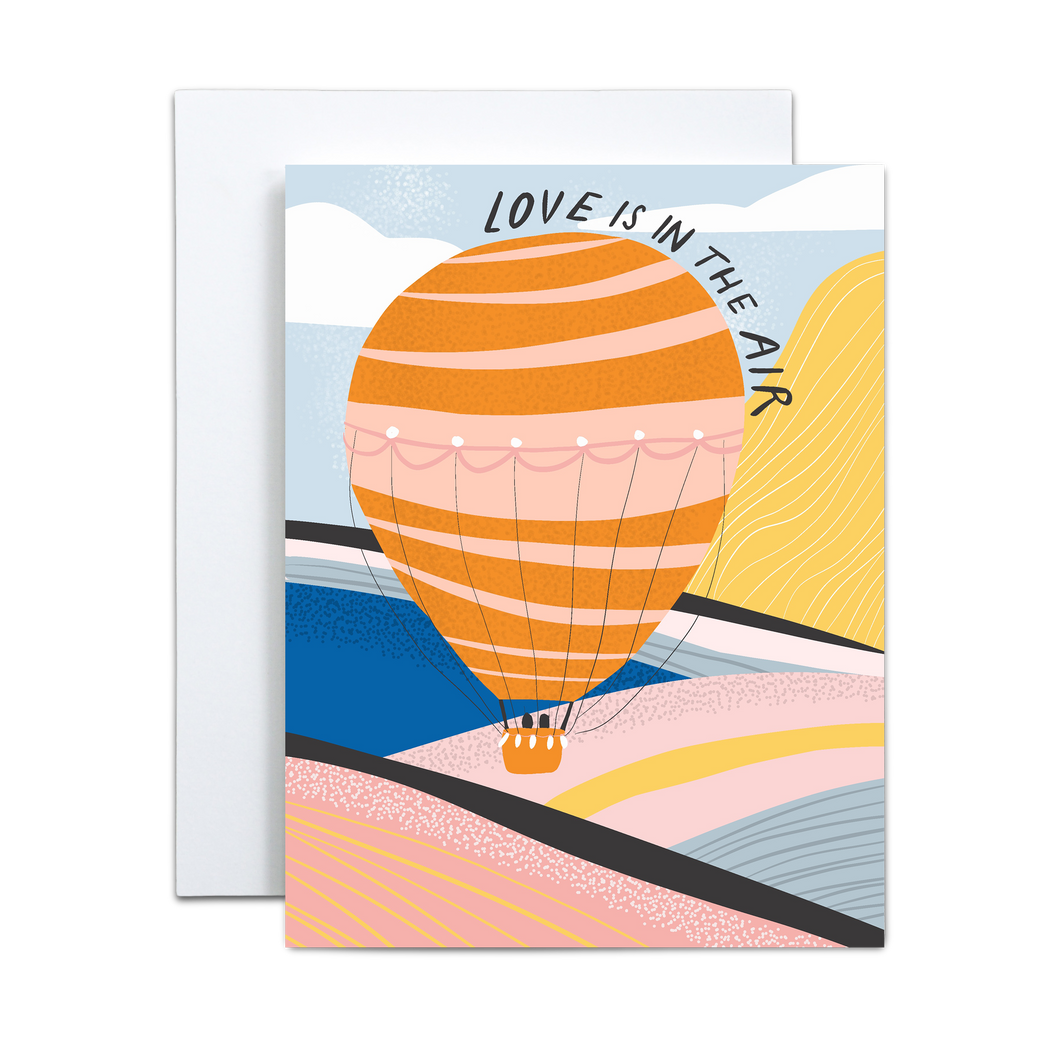 bold and abstract pink, yellow, cobalt, baby blue, black, and whitemountain and valley landscape with various textures and a pink and orange hot air ballon with two black shapes in the basket with 'love is in the air' text circling the balloon greeting card