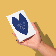 Load image into Gallery viewer, hand holding simple dark blue heart with &#39;sending you lots of love&#39; in white in the center on a white background greeting card
